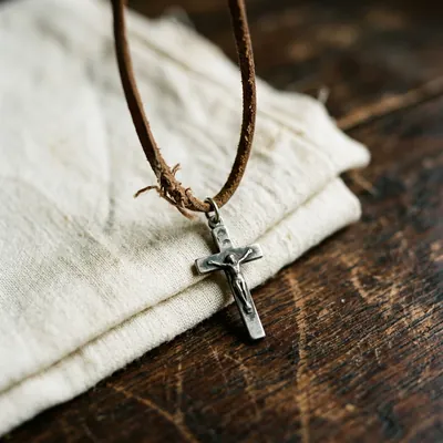 A small antique silver Catholic crucifix on a worn brown leather cord resting on folded cream linen · macro close-up with shallow depth of field